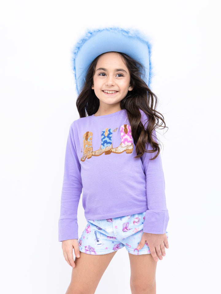 Young girl wearing a purple shirt with a cowboy boot design and western shorts, standing against a white background.
