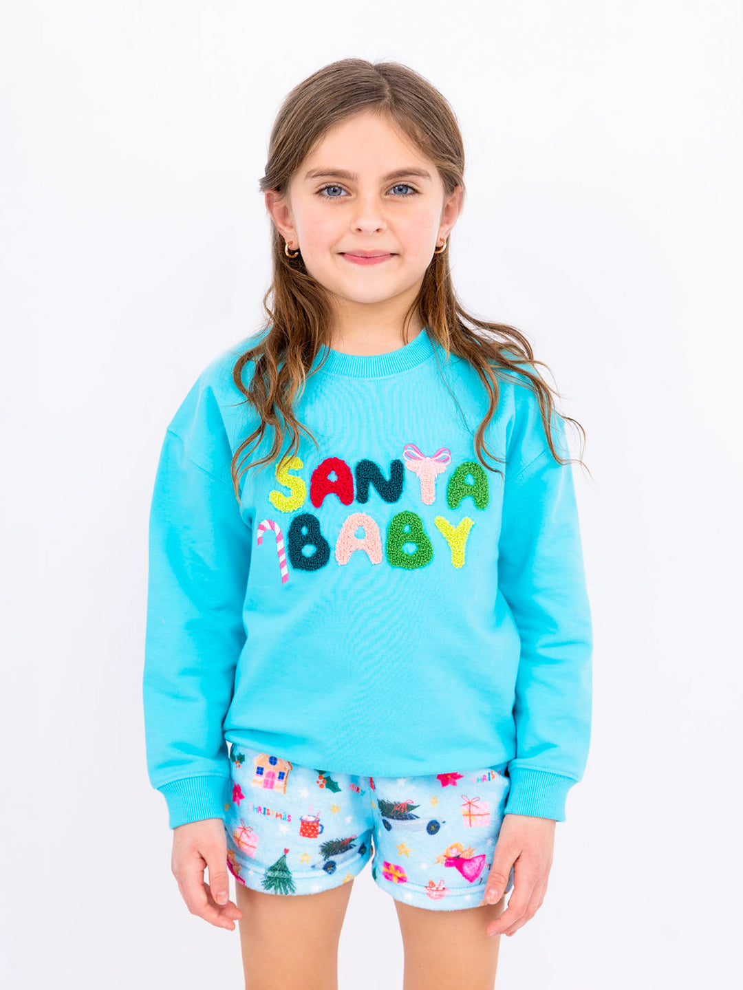 Young girl wearing a blue sweater with colorful Santa Baby logo and Christmas plush shorts on a white background