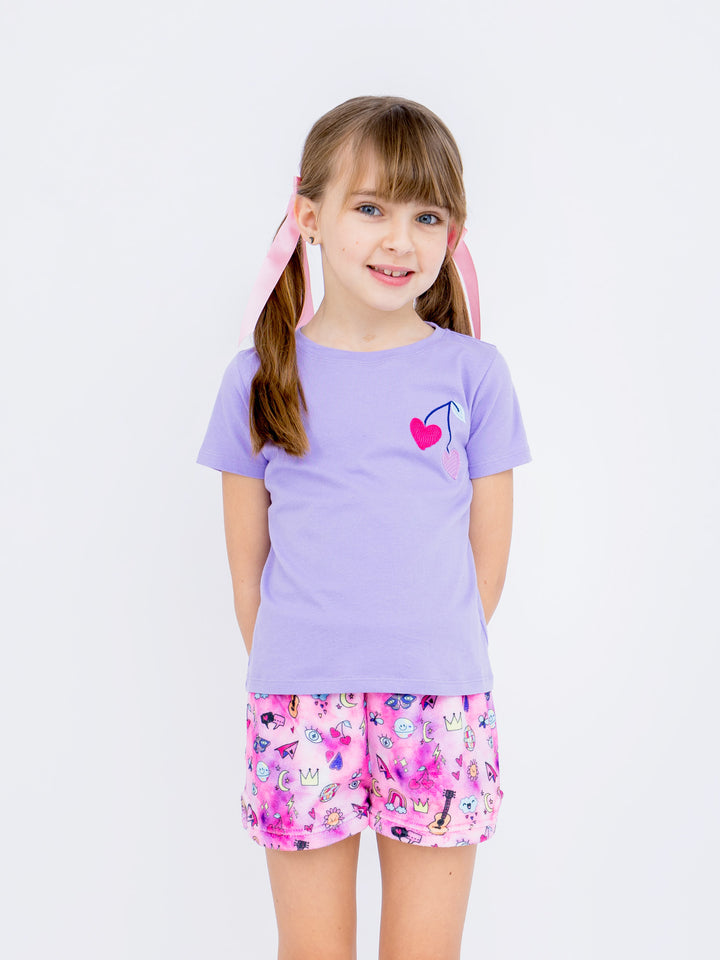 Young girl wearing a purple shirt with a cherry design and patterned shorts on a white background