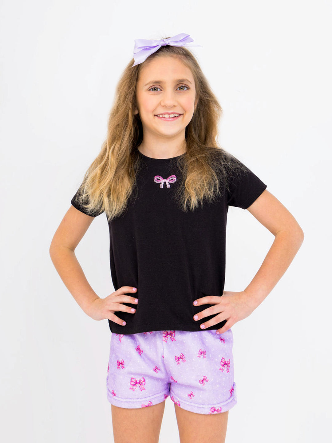 Young girl wearing a black t-shirt with a pink bow and purple shorts with pink bows on a white background