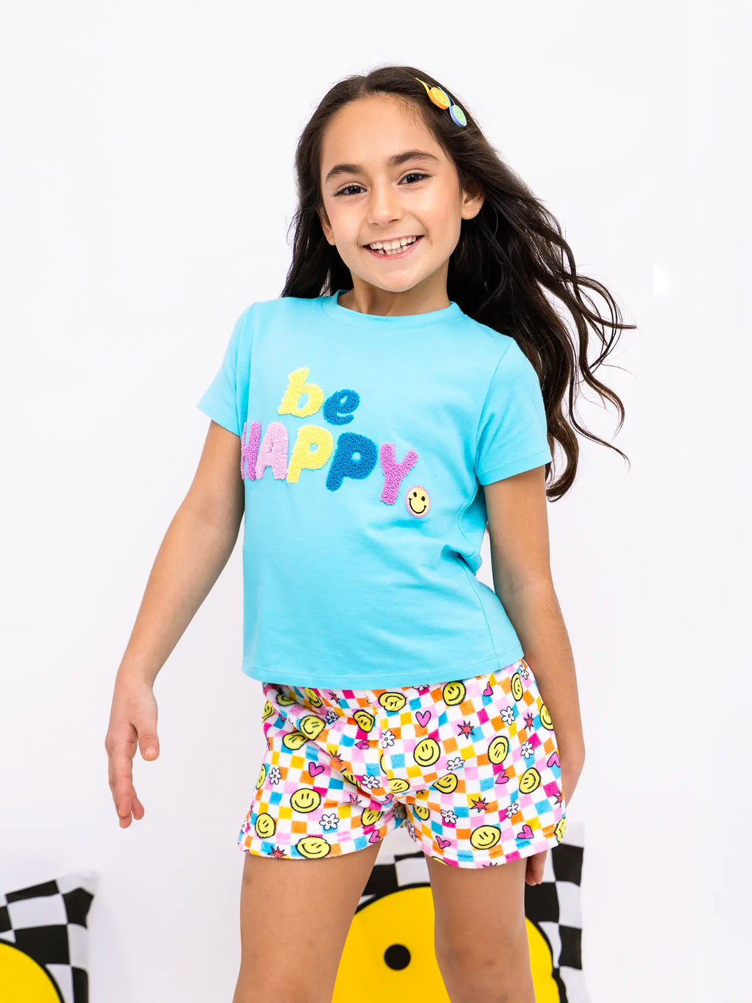 Young girl wearing a 'be happy' t-shirt and colorful smiley checker printed shorts on a white background