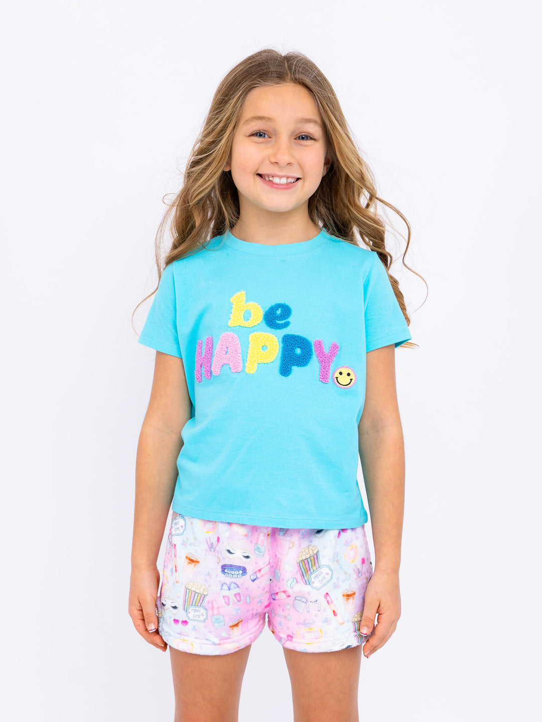 Young girl wearing a blue 'be happy' t-shirt and sleepover printed plush shorts on a white background