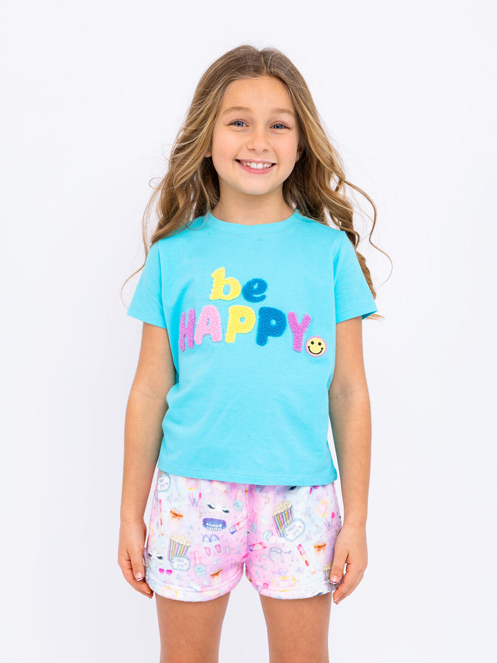 Young girl wearing a blue 'be happy' t-shirt and sleepover printed plush shorts on a white background