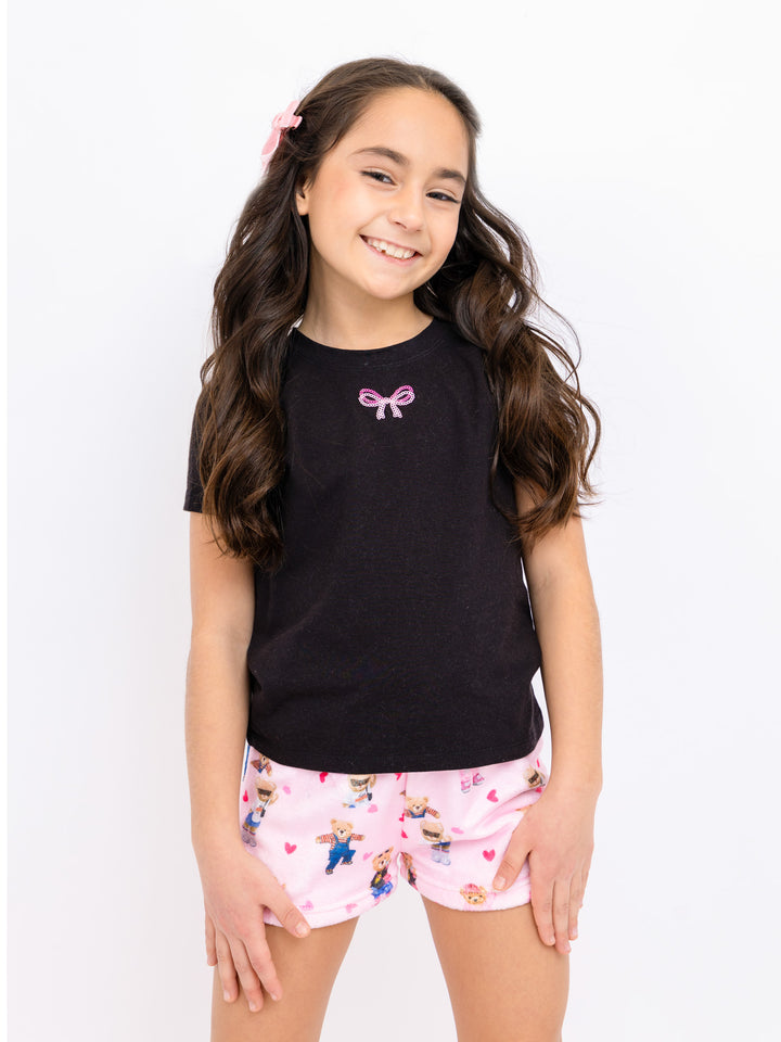Young girl wearing a black top with a pink bow and pink shorts with teddy print on a white background