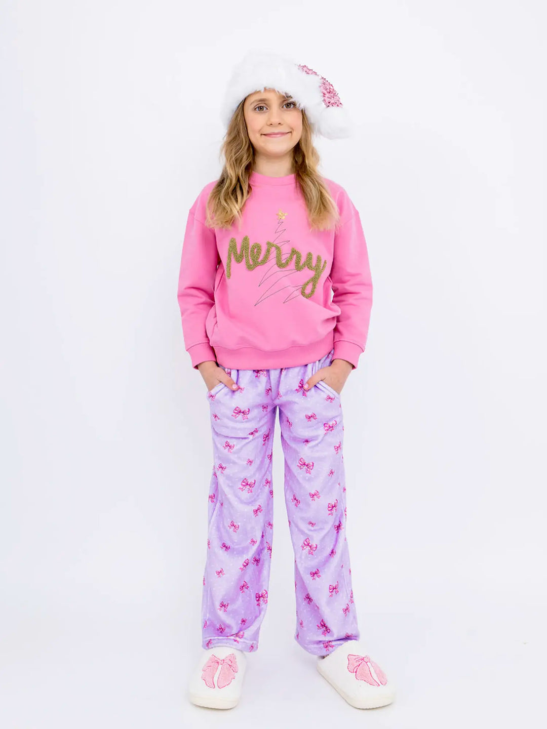 Young girl wearing a pink 'Merry' sweater and purple pajama loungewear plush pants with a white background