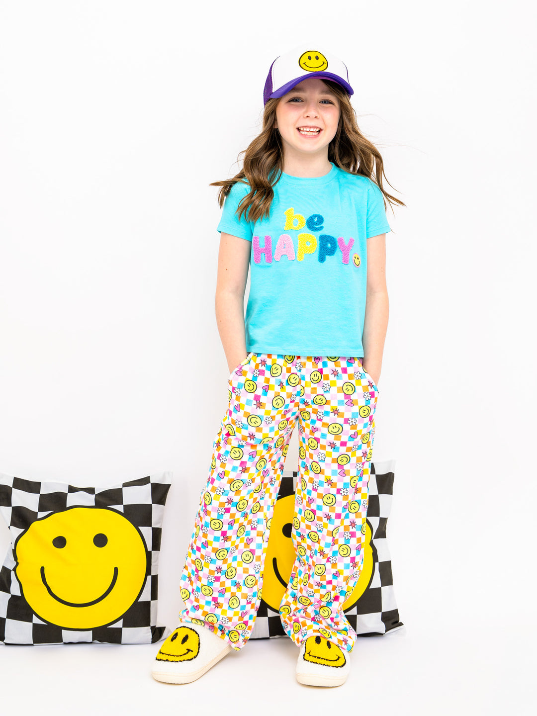 Child wearing a 'be happy' t-shirt and colorful pants with smiley face design, standing against a white background.