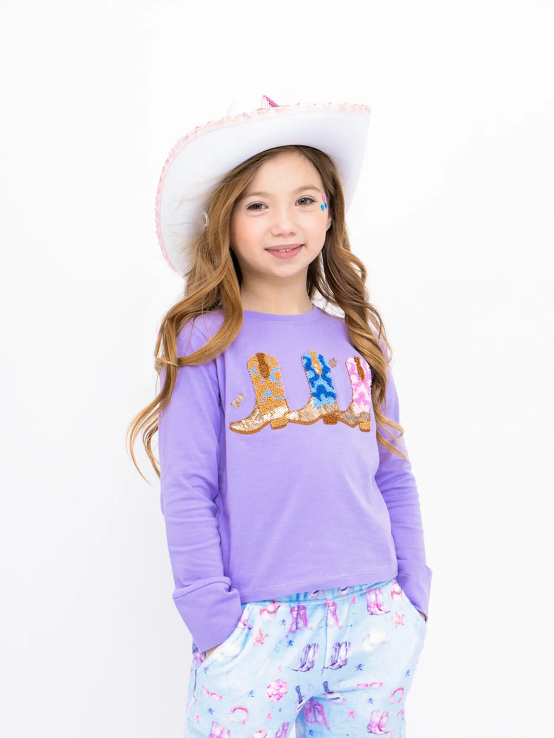 Young girl wearing a purple shirt with cowboy boots and a white hat on a white background