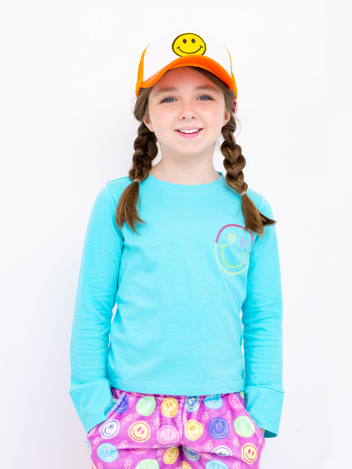 Young girl wearing a blue long-sleeve shirt with a smiley face design, colorful pants, and an orange cap with a smiley face on a white background.