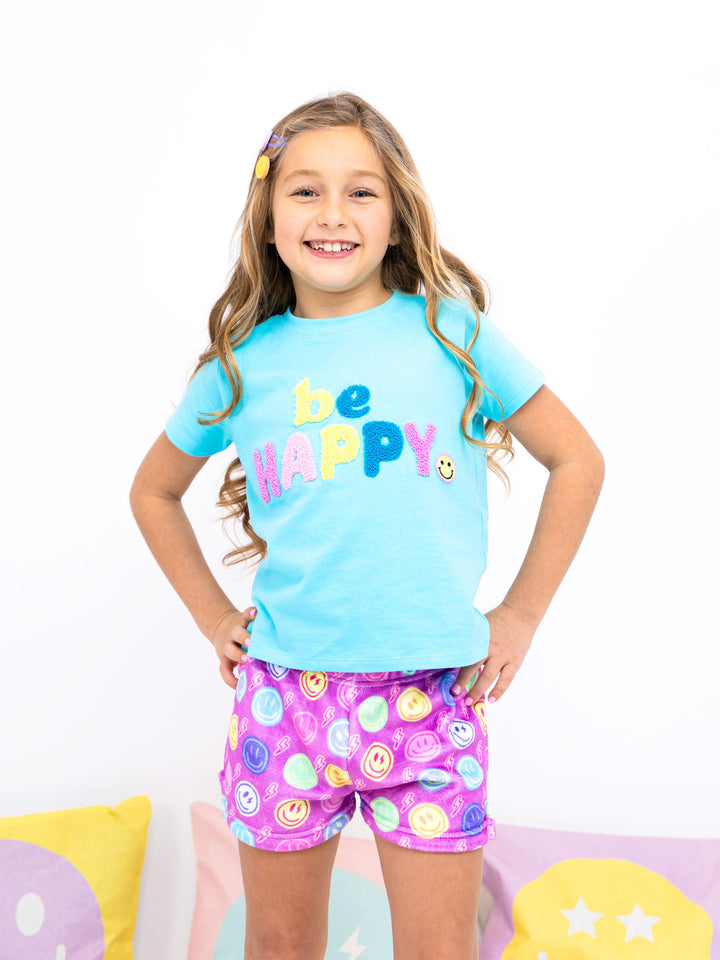 Young girl wearing a blue 'be happy' t-shirt and colorful shorts on a white background