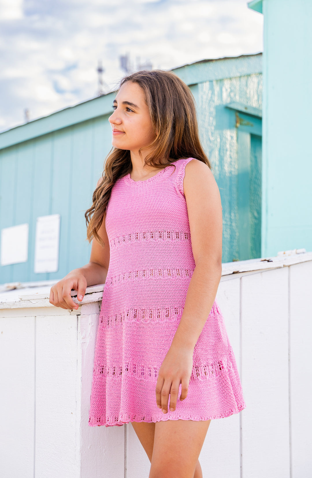 Tween girl wearing a pink crochet dress standing by a white railing with a blue building in the background