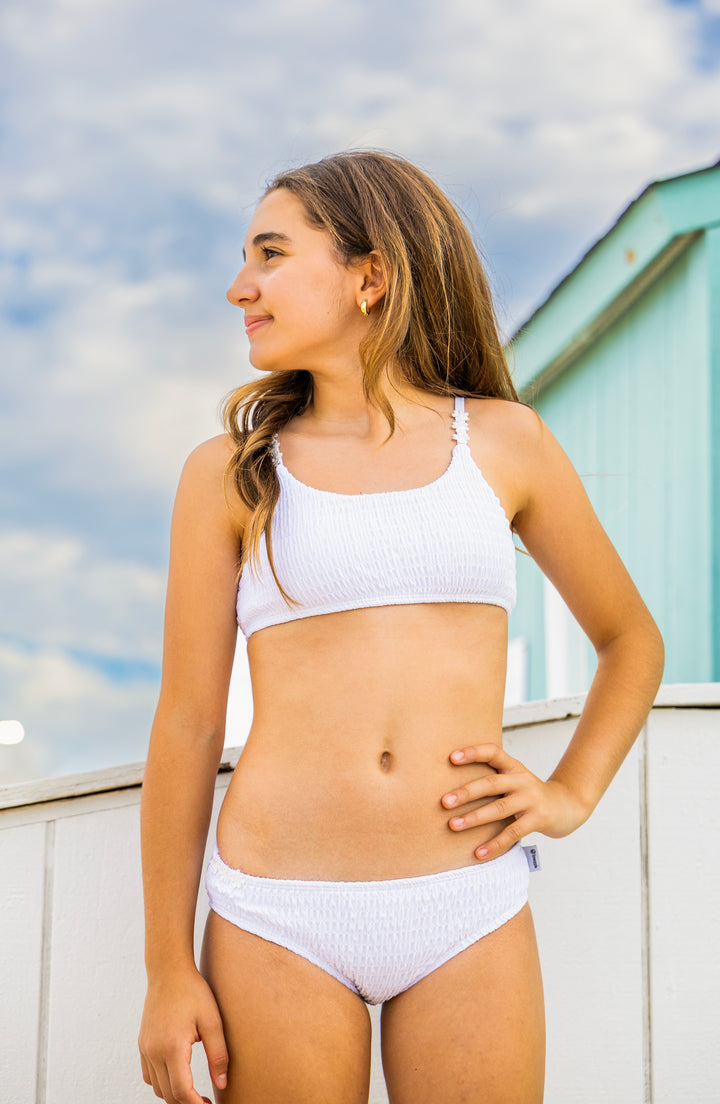 Tween girl wearing a white bikini standing outdoors with a blue sky and building in the background