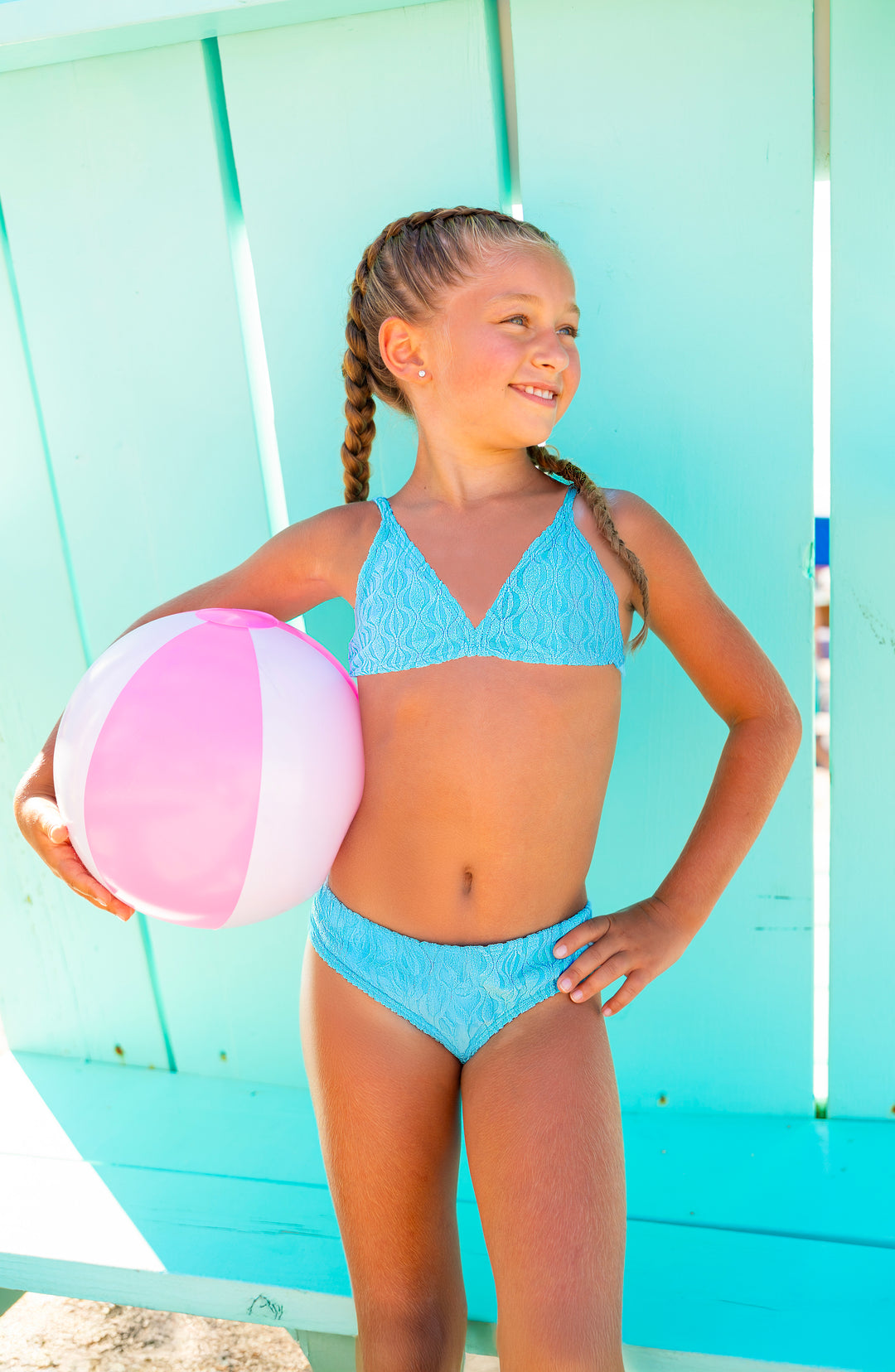 Young girl in a blue bikini holding a pink and white beach ball against a light blue background.