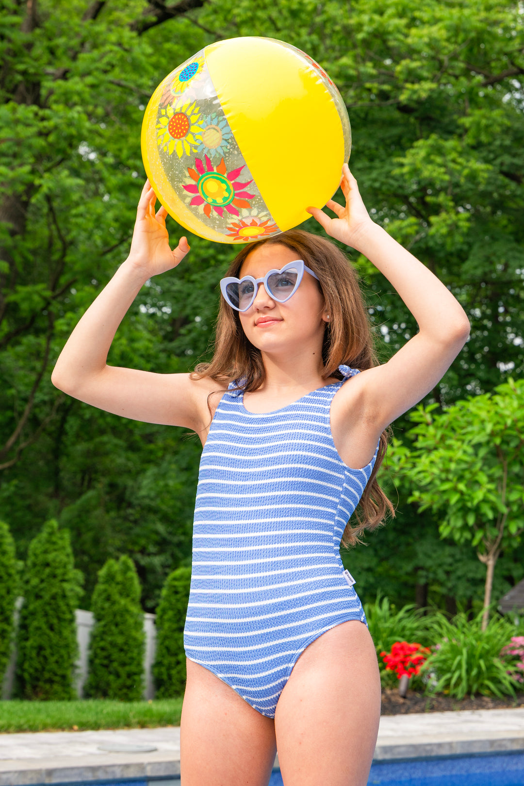 girl in a blue striped one piece swimsuit holding a colorful beach ball outdoors.
